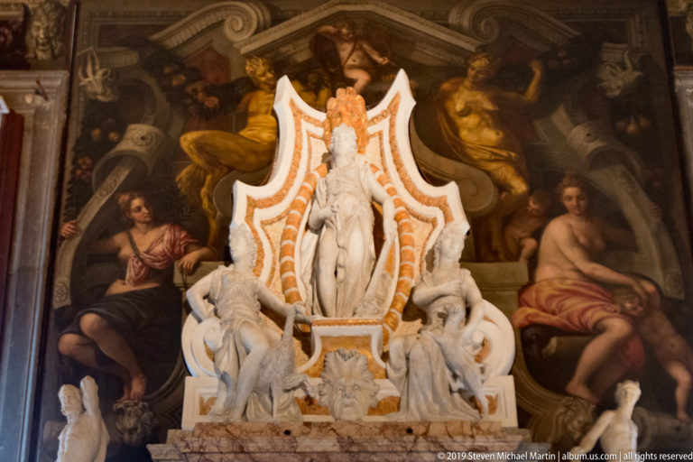 White marble monument with a standing central figure and surrounding sculptures placed before a large dark wall painting.