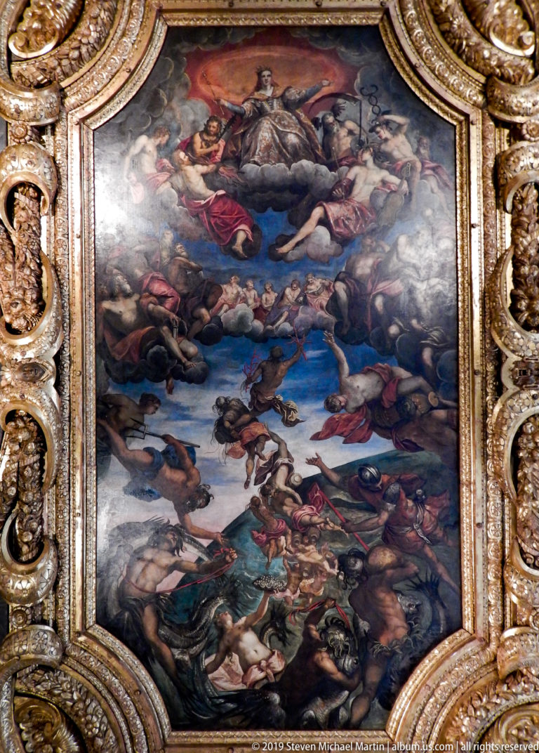 Large allegorical ceiling painting crowded with figures, set within a deeply carved gilded frame and viewed from directly below.