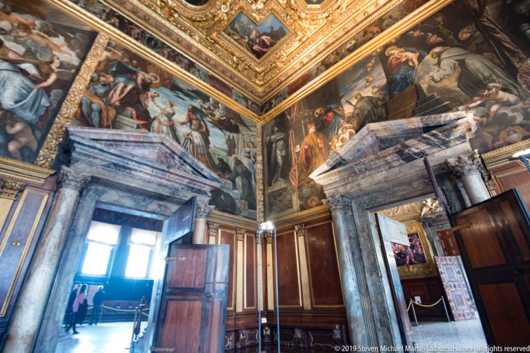 Corner of an ornate chamber with large wall paintings, dark wood paneling, massive gray stone doorways, and open passages.