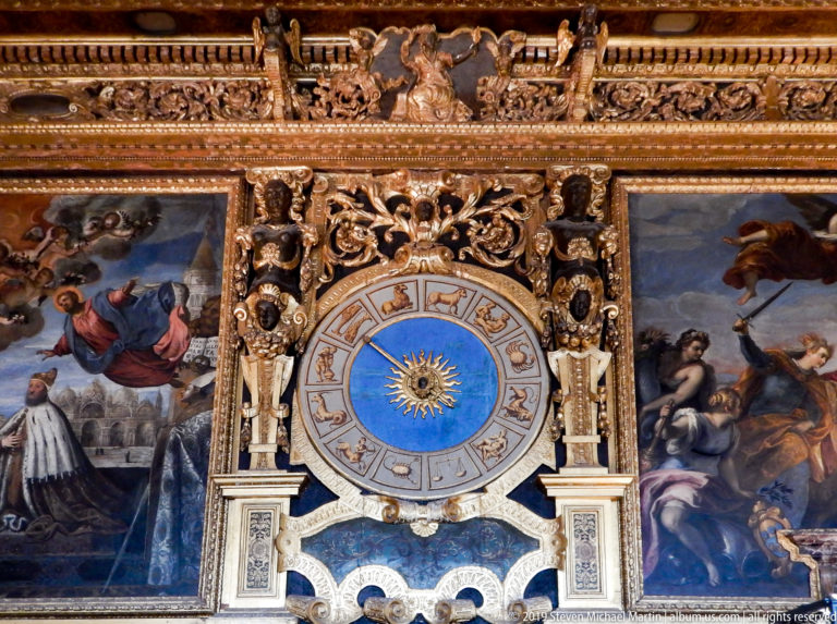 Blue zodiac clock centered in gilded wall decoration between large painted panels with allegorical figures.