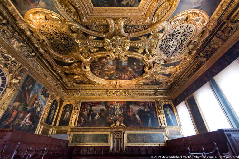 Diagonal view of ceremonial room with gilded ceiling, large wall painting, lower friezes, wooden seating, and bright windows.