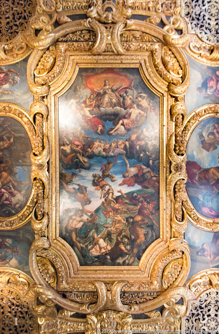 Rectangular multi-figure ceiling painting surrounded by thick gilded scrollwork, with smaller painted compartments at the sides.