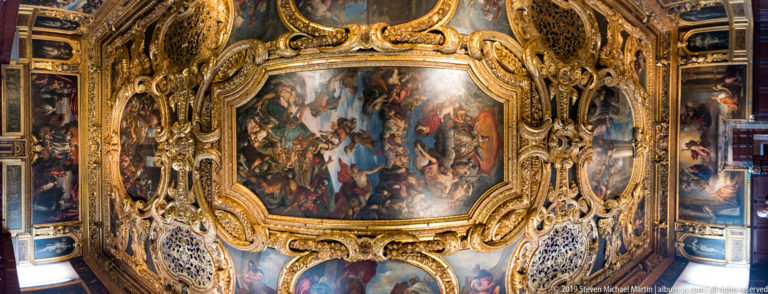 Angled view of a gilded ceiling with a large central narrative painting, smaller scenes, scrollwork, and pierced panels.