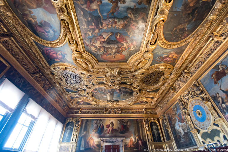 Corner of ornate palace room with gilded painted ceiling, tall window, wall paintings, and blue zodiac clock.