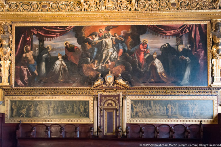 Ceremonial wall with a large allegorical painting above two long monochrome scenes, central doorway, and wooden seating.