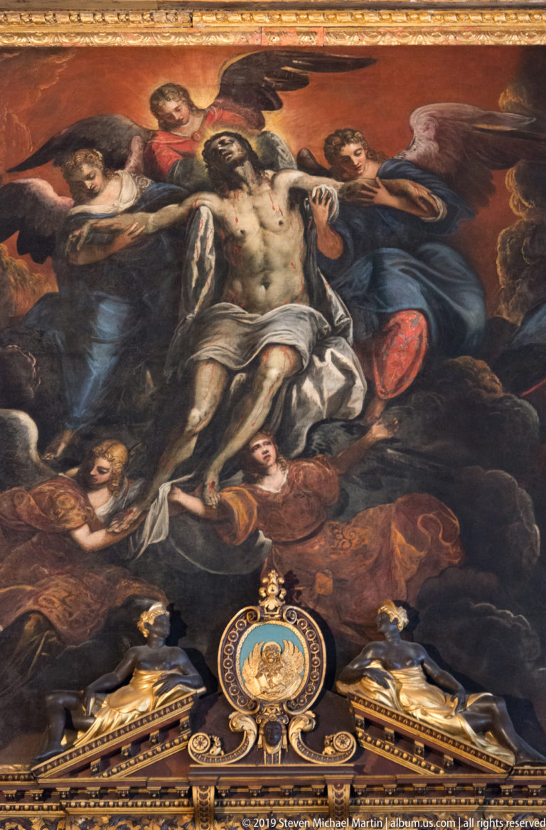 Pale body of Christ supported by mourners in a dark religious painting above ornate gilded decoration.