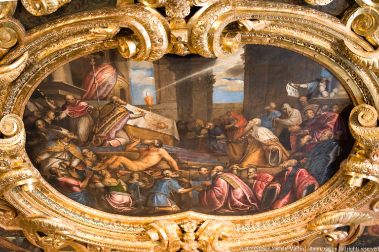 Oval narrative painting of many figures in an interior, enclosed by heavy gilded scrollwork and carved ornament.