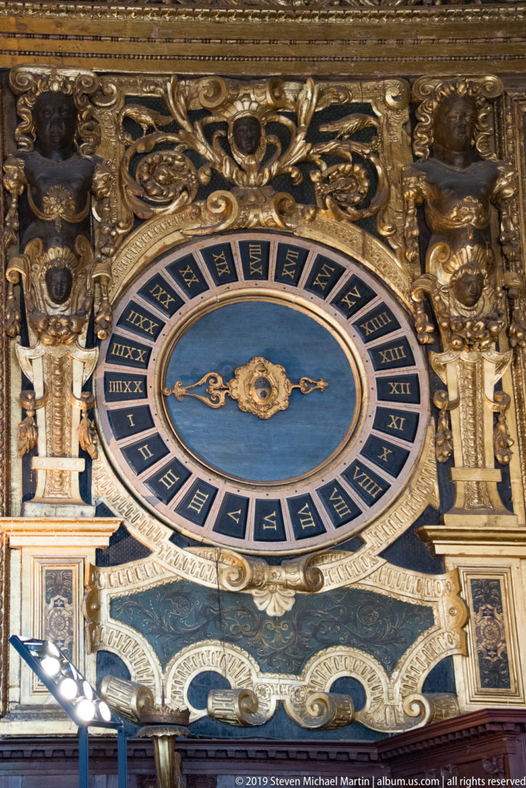 Blue circular twenty-four-hour clock dial with Roman numerals set in elaborate gilded carved decoration.