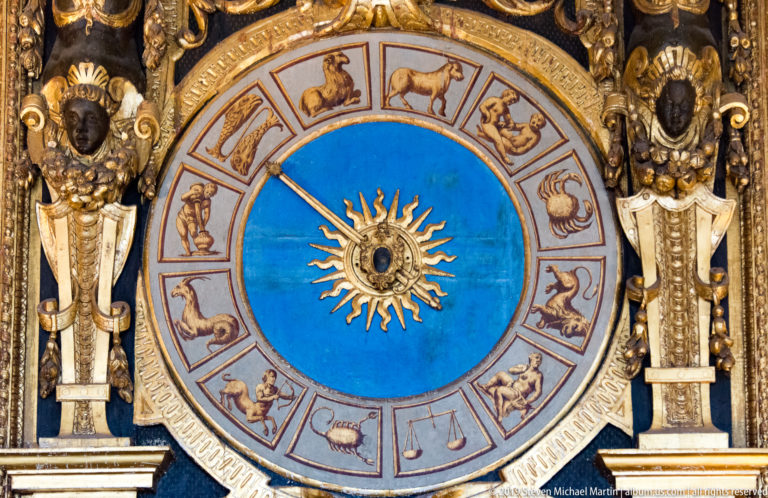 Blue zodiac clock dial with twelve signs around a gilded sunburst, framed by sculpted figures and gold ornament.