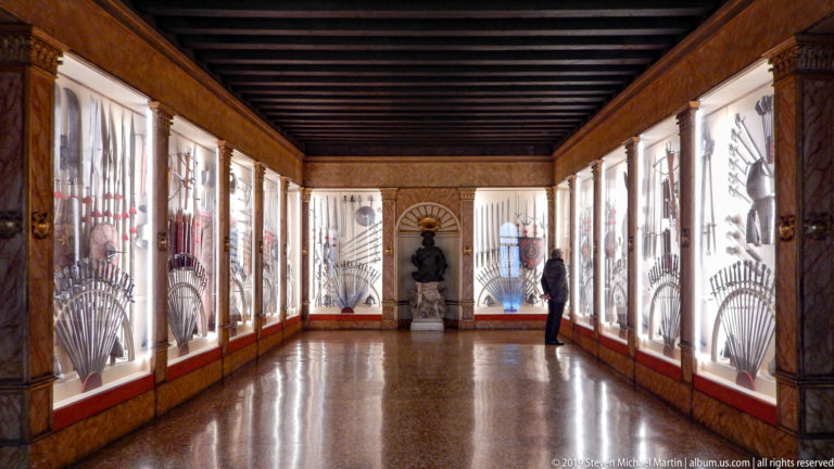 Long palace armory gallery with illuminated weapon cases on both walls, dark ceiling beams, and a visitor near the far end.