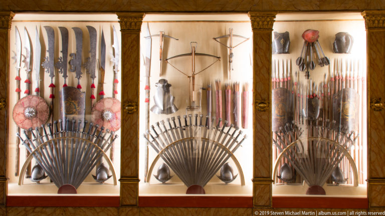 Three illuminated armory cases filled with fan-shaped arrangements of polearms, guns, bows, blades, shields, and armor.
