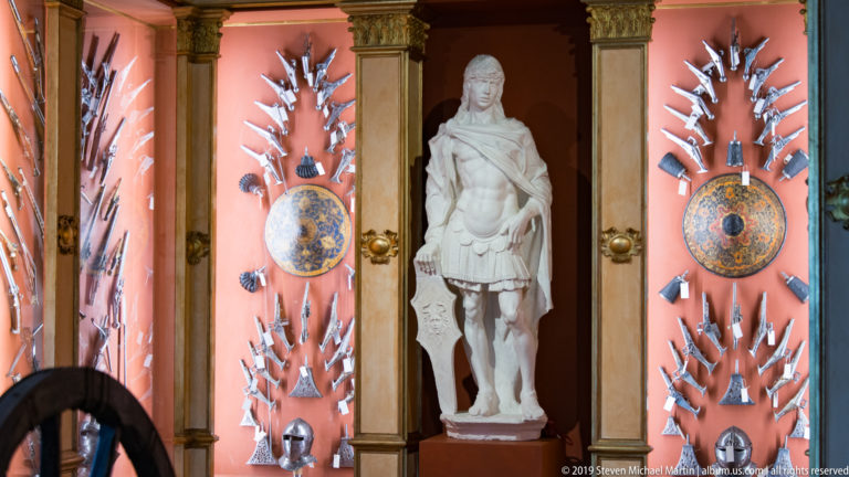 White warrior statue in a niche between symmetrical displays of weapons, armor pieces, and painted shields.