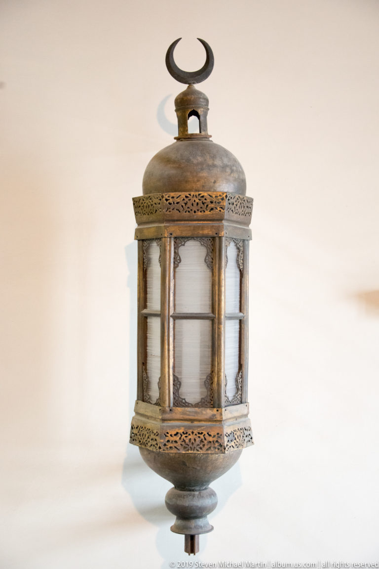 Small metal lantern with translucent cylindrical body, pierced bands, slender supports, and crescent-shaped finial.