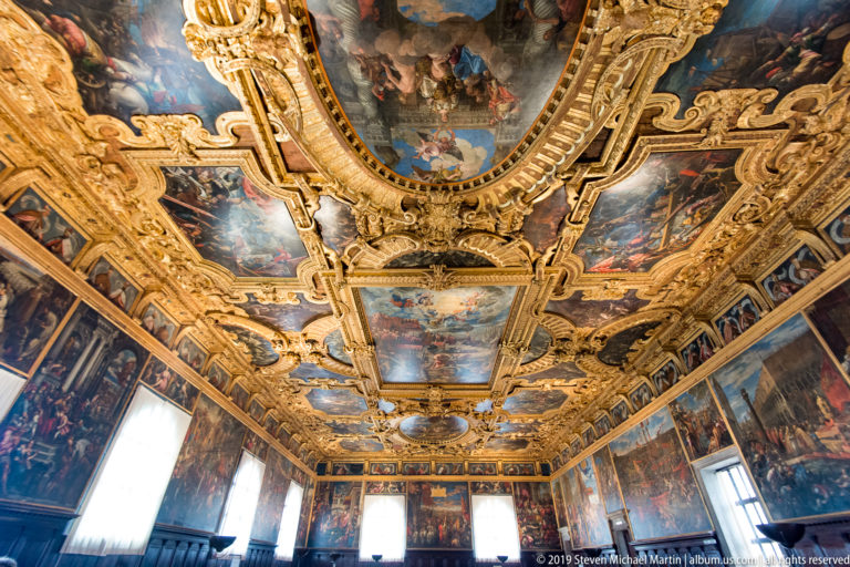 Wide interior view beneath a gilded painted ceiling, with large wall paintings and bright windows at the far end.