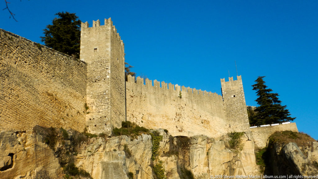 Long crenellated stone wall with square towers rising from a rocky Monte Titano slope beneath a deep blue sky.