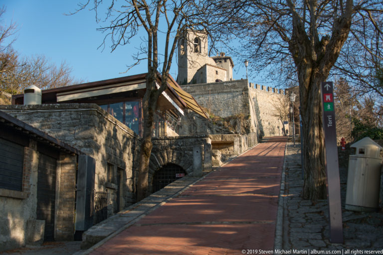 Red pedestrian ramp climbing between stone walls and bare trees toward a crenellated tower and fortification.