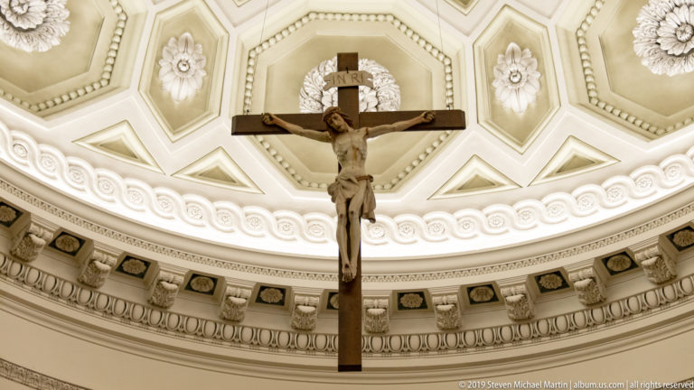 Large crucifix with sculpted Christ centered beneath an ornate cream-colored dome with coffering, rosettes, and classical moldings.