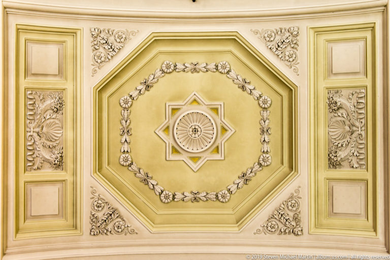 Ornate cream-and-gold octagonal ceiling panel with floral garlands, rosettes, geometric moldings, and a star-shaped central feature.