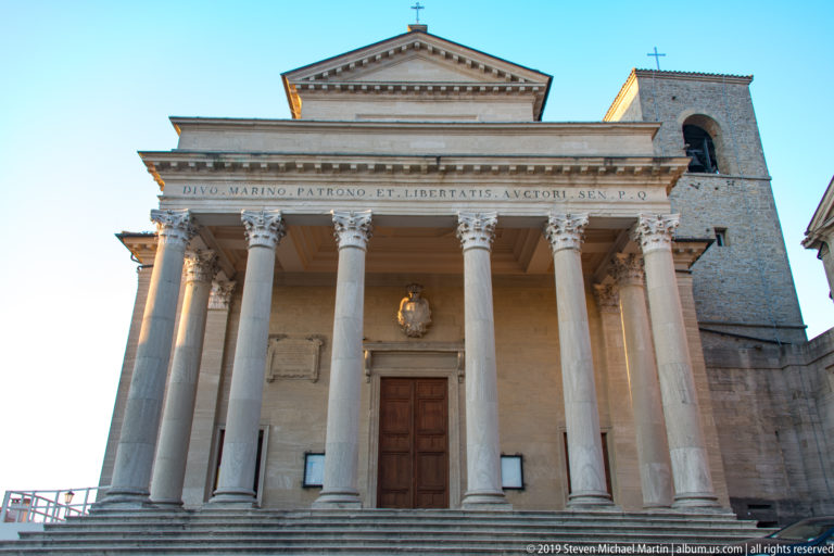 Front of Basilica di San Marino with Corinthian columns, triangular pediment, broad steps, inscription, and bell tower at right.
