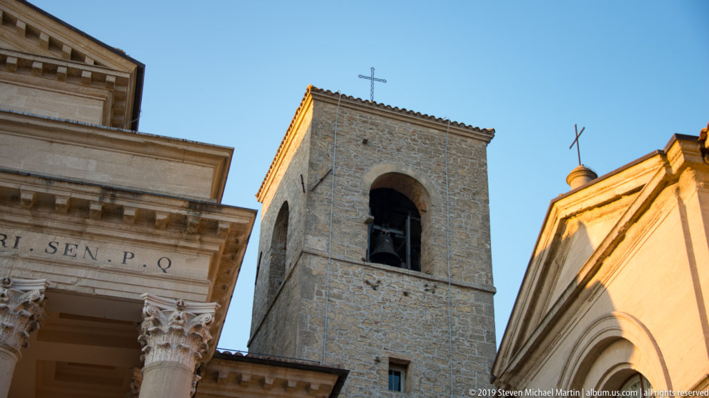 Basilica di San Marino stone bell tower with large arched bell opening and cross between adjoining church façades.
