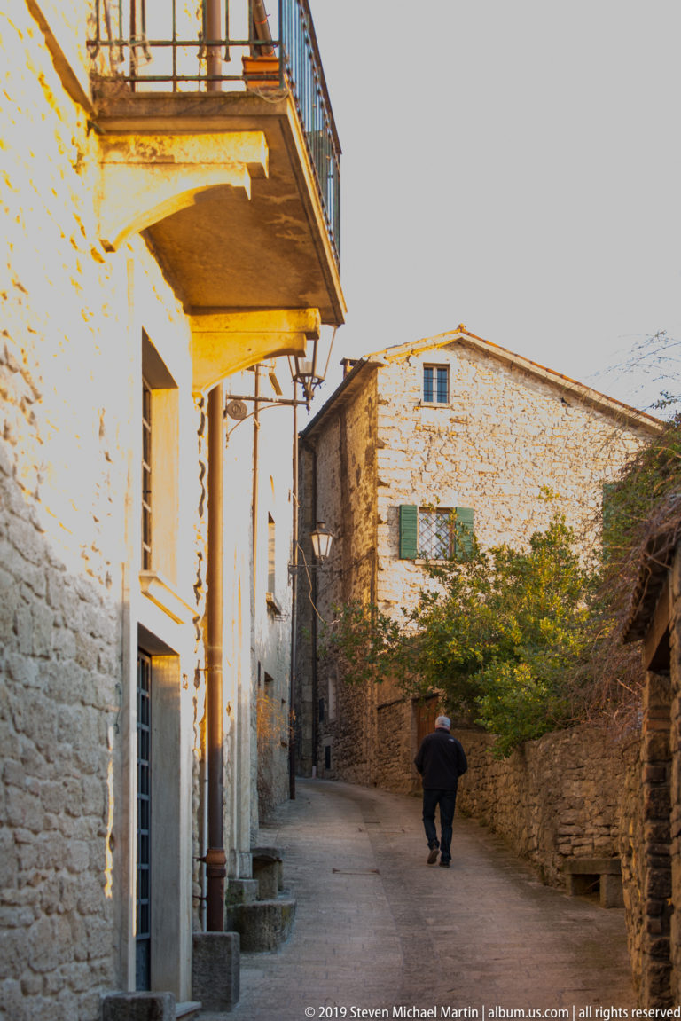 Quiet stone lane between masonry buildings with balconies and walls, where one pedestrian walks away toward a sunlit house.