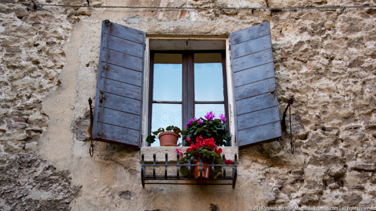 Open gray wooden shutters around a dark-framed window with red and purple potted flowers on a small iron rail.