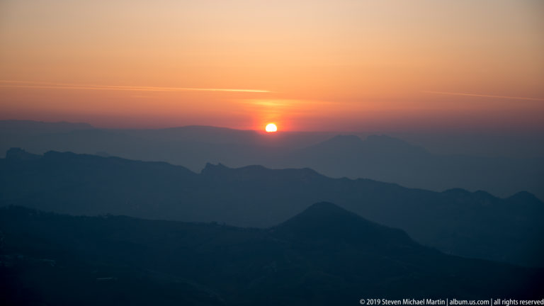 Bright orange sun above layered dark mountain ridges beneath a peach-colored evening sky.