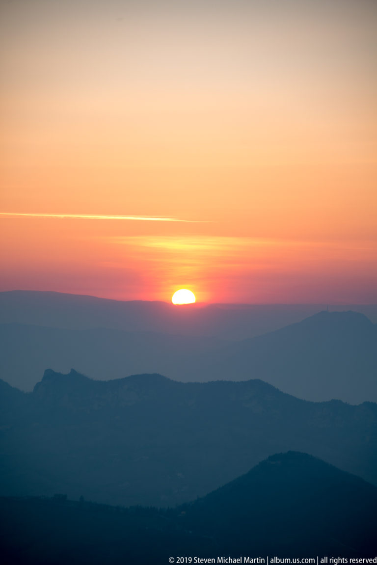 Sun setting behind layered blue mountain ridges beneath an orange-pink sky and thin illuminated cloud.