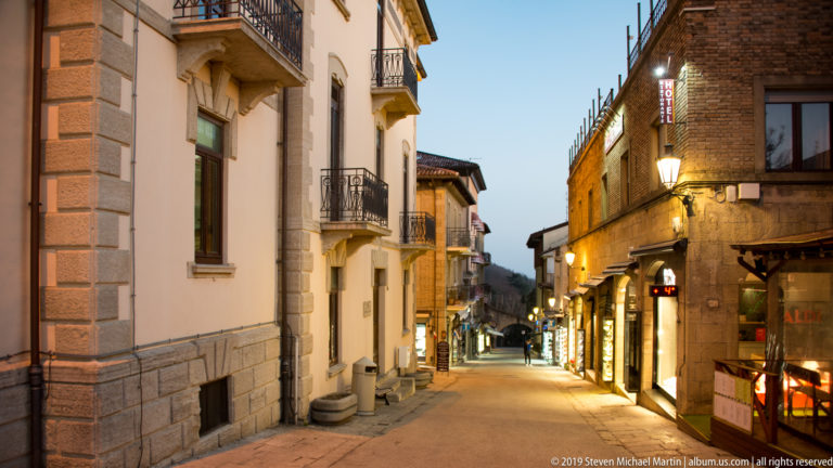 Twilight street descending between pale buildings with balconies and warmly lit shops along the right side.