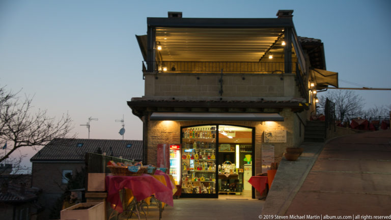 Small brightly lit shop beneath a roof terrace and modern canopy, with outdoor tables on a steep hillside.