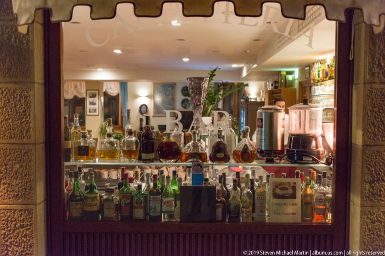 Bar window filled with liquor bottles, glassware, coffee equipment, and large BAR lettering within a stone surround.