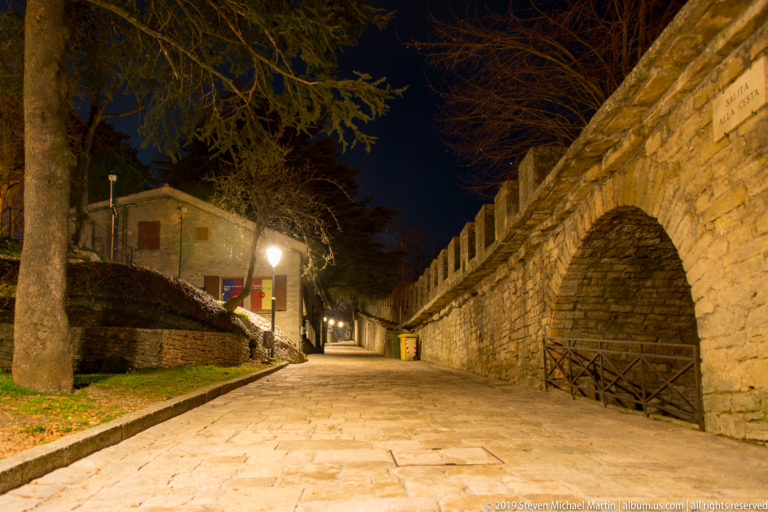 Stone-paved lane at night beside a high crenellated wall with a large recessed arch, railing, trees, and streetlamps.
