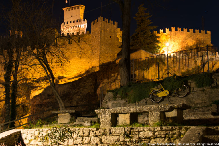 Golden-lit crenellated fortress walls and square tower above a terraced slope, railing, trees, and a parked bicycle.