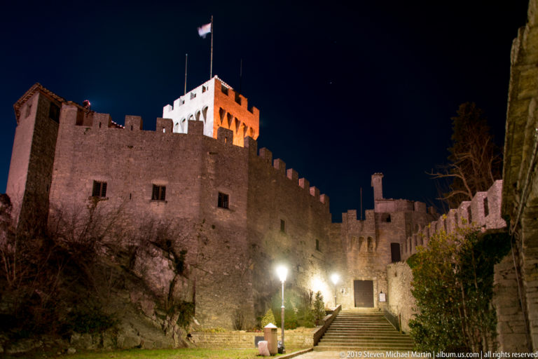 Floodlit fortress walls and square tower with San Marino flag above a doorway and stair in a small inner approach.