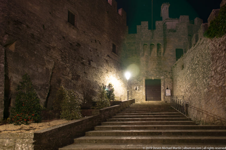 Wide stone stair between high fortress walls leading to a lit rectangular doorway beneath crenellations at night.