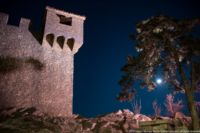 Low-angle floodlit stone tower with deep projecting corbels, rocky base, pine tree, and bright moon in a dark sky.