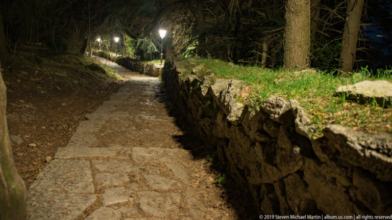 Stone-paved woodland path at night beside a low rough-rock wall, trees, grass, and a glowing path lamp.