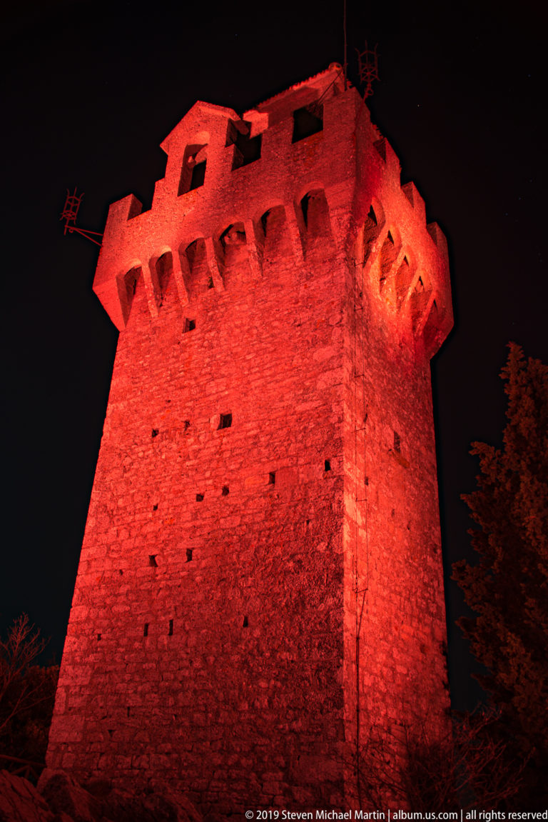 Low-angle view of a massive square stone fortress tower illuminated red, with projecting corbels and small wall openings.