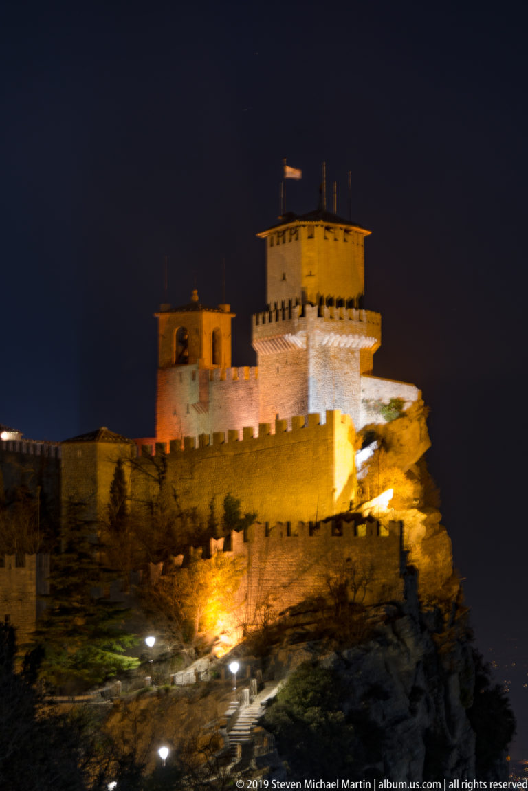 Illuminated Second Tower, Cesta or Fratta, with central keep, crenellated walls, and rocky summit against a black sky.
