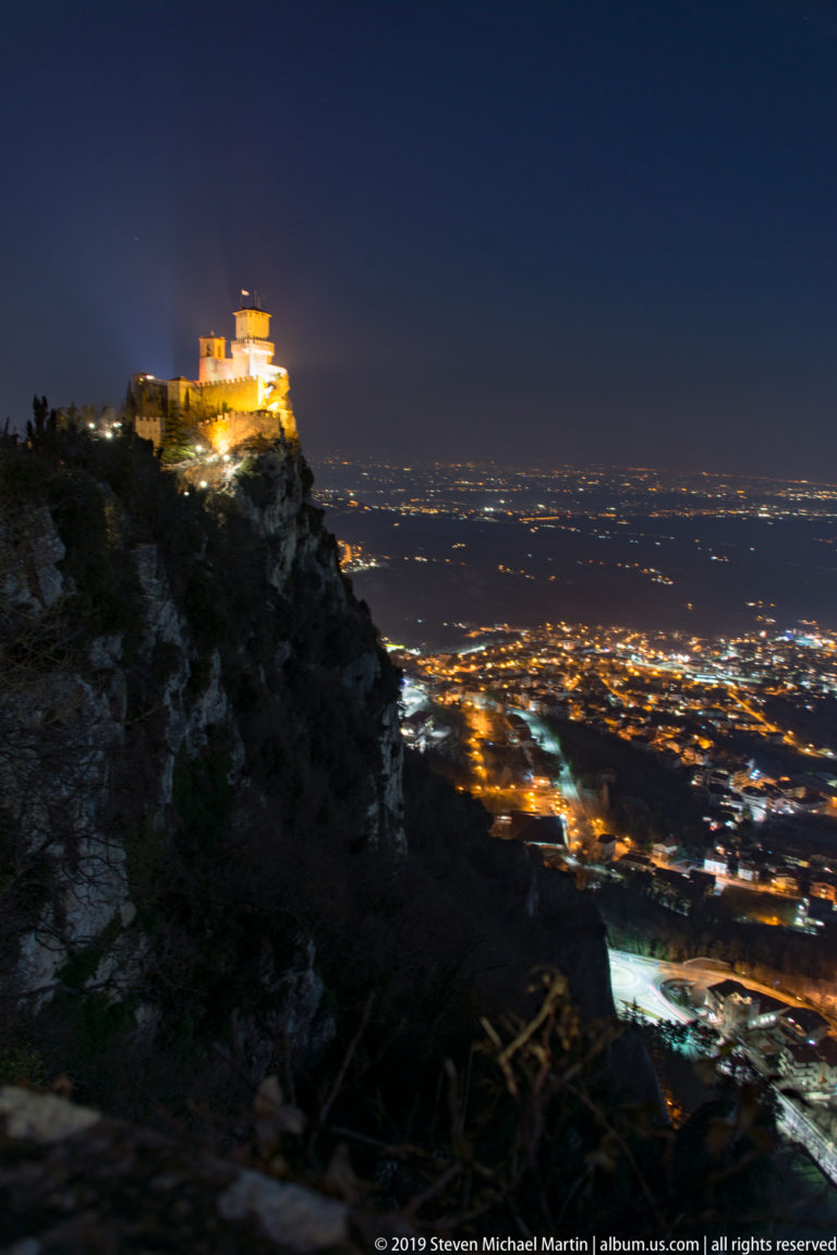Floodlit fortress on a rocky cliff at left overlooking a broad field of city lights beneath a deep blue evening sky.