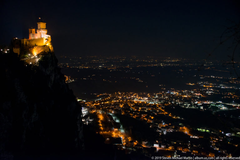 Illuminated hilltop fortress at far left above a vast nighttime plain filled with city and road lights.