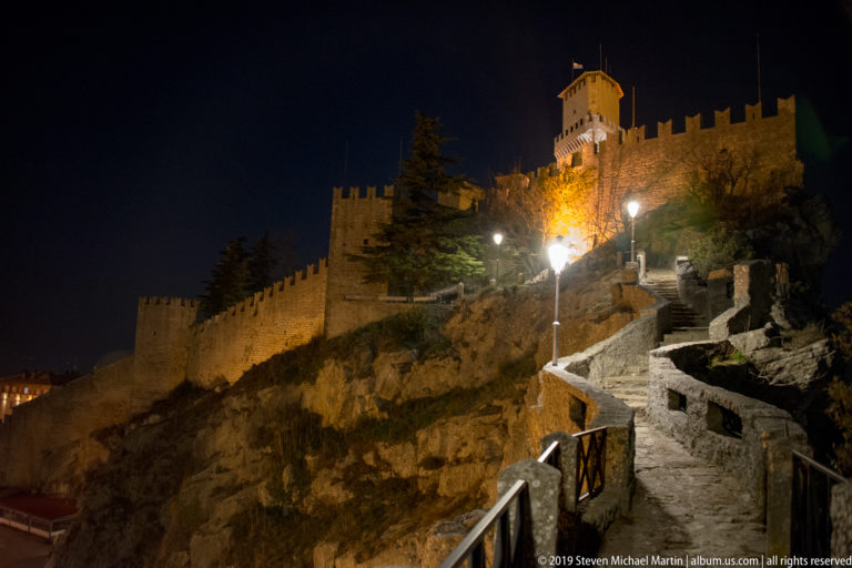 Stone path and steps with modern lamps climbing over rock toward illuminated crenellated walls and a square fortress tower.