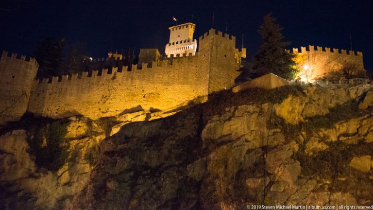 Golden floodlit crenellated walls and towers rising above a rugged Monte Titano rock face at night.