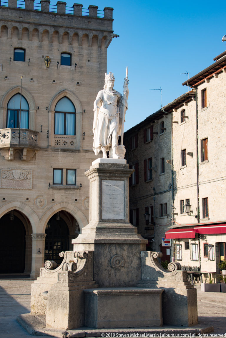 White marble Statue of Liberty on a tall pedestal in front of Palazzo Pubblico’s arched façade in San Marino.
