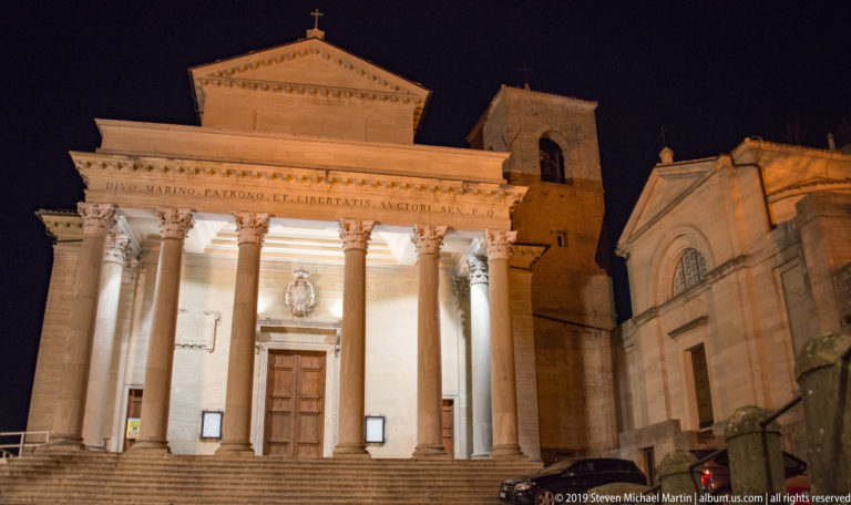 Illuminated Basilica di San Marino with Corinthian portico, broad steps, pediment inscription, bell tower, and adjoining chapel.