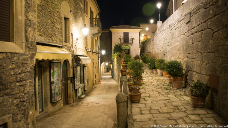 Narrow stone lane at night with shop awnings, streetlamps, terracotta pots, and a high masonry wall.