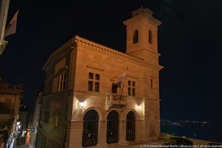 Illuminated stone civic building with arched lower windows, balcony, and square bell tower against a dark sky.