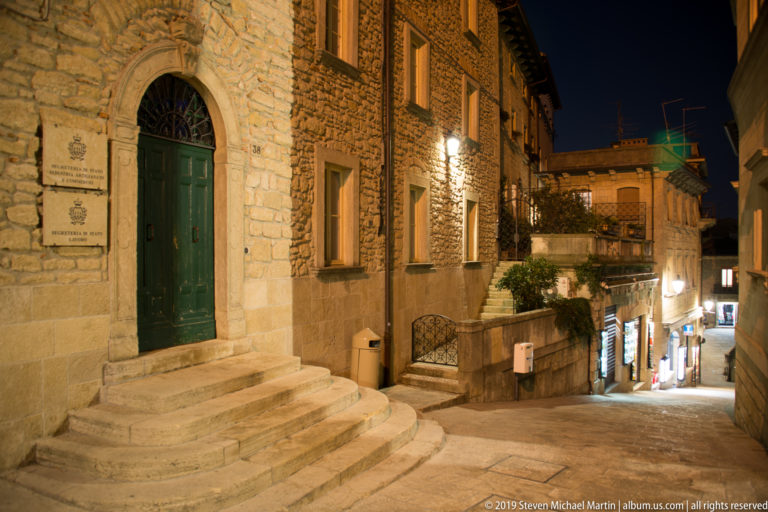 Green double door above curved stone steps on a masonry building beside a steeply descending illuminated street.