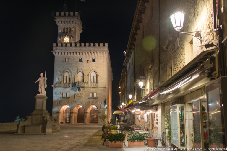 Night streetscape with Palazzo Pubblico ahead, Statue of Liberty at left, and awnings and café seating along the right.