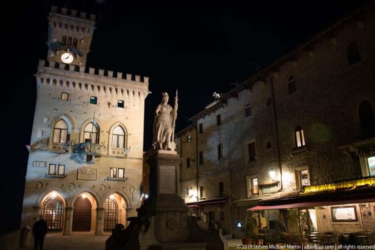 Night view across Piazza della Libertà with the Statue of Liberty, illuminated Palazzo Pubblico, and stone buildings with restaurants.